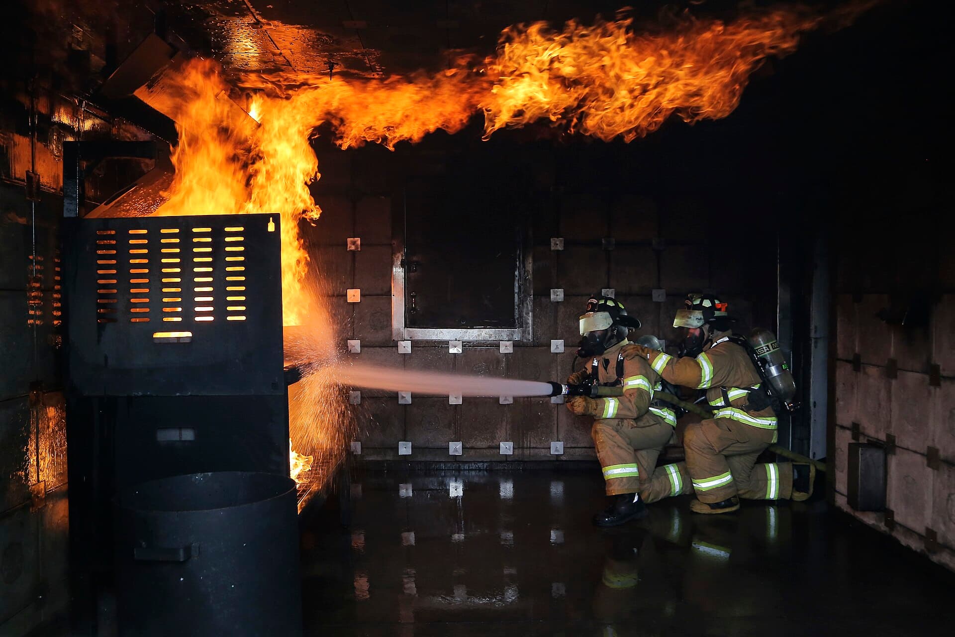Firefighters in full gear driving a hose into roaring flames